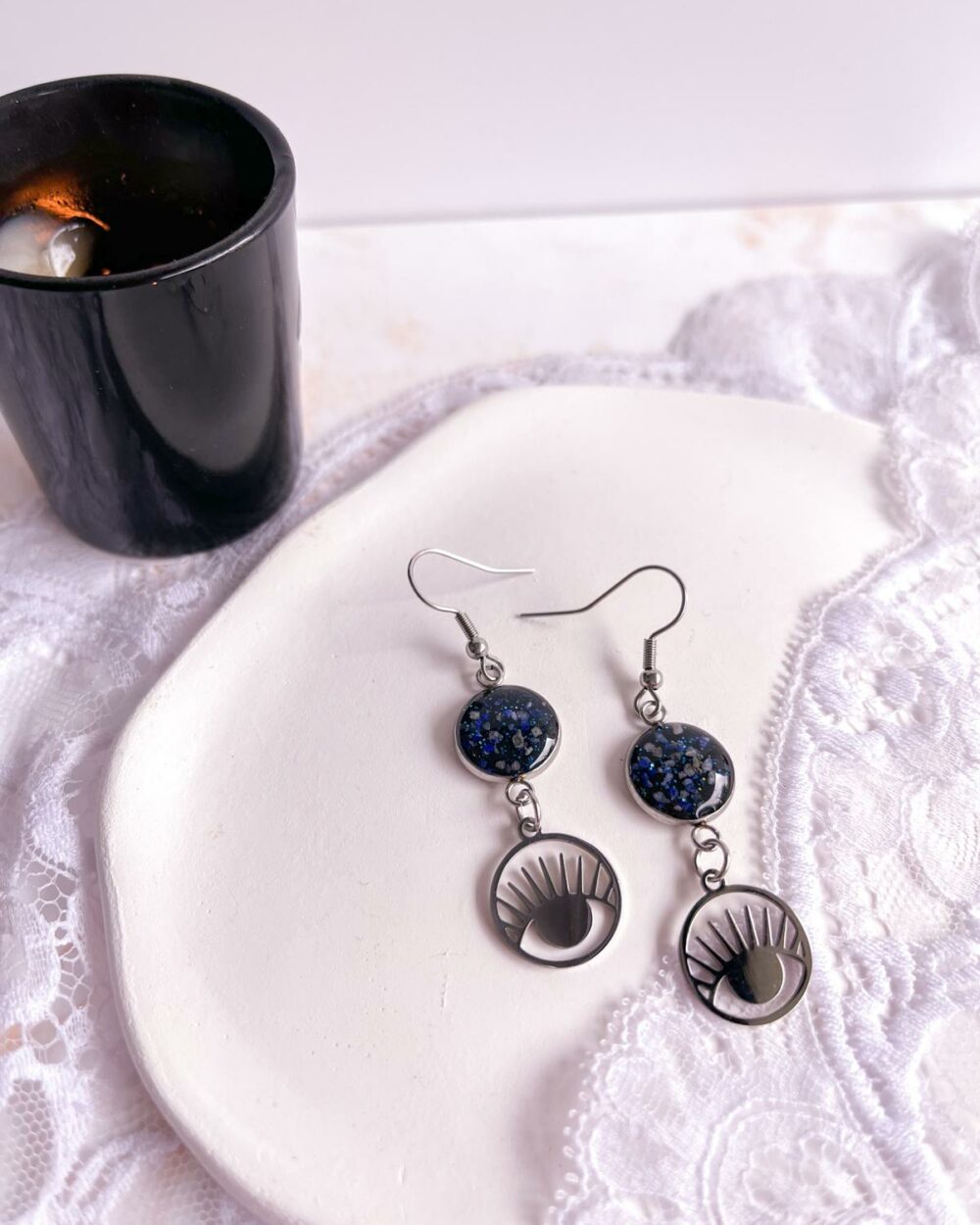 Boucles argentées en acier inoxydable avec médaillon noir de 12 mm incrusté de sable de lapis-lazuli et breloque œil dans rond ajouré.